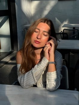 A woman with long hair sits at a table in a bright cafe, enjoying a moment of calmness while sunlight filters through the window.