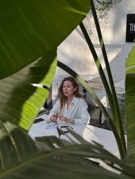 A woman sits at a table in a bright, modern workspace, focused on her tasks amid vibrant green plants.
