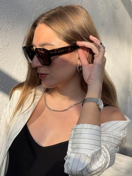 A woman with long hair wearing stylish sunglasses adjusts her eyewear while enjoying a sunny day outdoors.