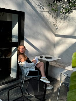 A woman sits comfortably at a small table in a bright cafe, enjoying the sunshine and reading.