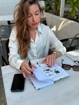 Woman sits at a cafe table, jotting notes in a notebook while enjoying the warm sunlight in a relaxed atmosphere.
