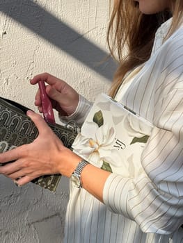 A woman is holding a patterned pouch and a floral notebook, showcasing her fashion sense against a sunlit wall.