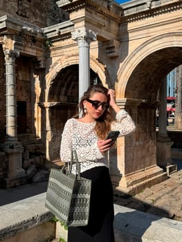 A young woman stands by historic stone ruins, checking her smartphone while wearing casual, stylish attire.