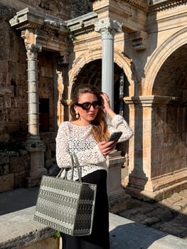 A woman dressed in a stylish outfit stands among ancient stone structures while checking her phone, enjoying a sunny day.