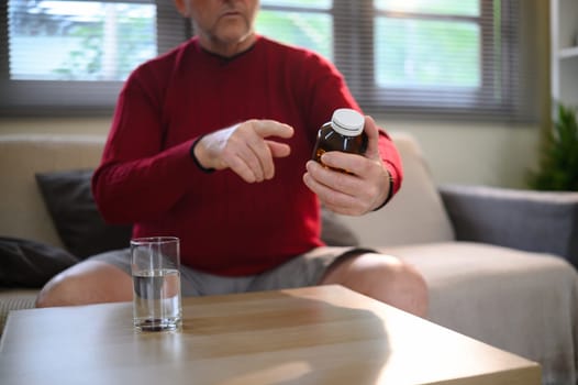 Mature man examining a prescription bottle while seated at home with a glass of water.