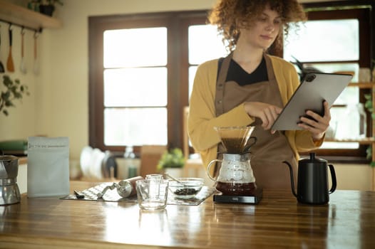Young woman uses digital tablet while preparing pour over coffee in a stylish kitchen.