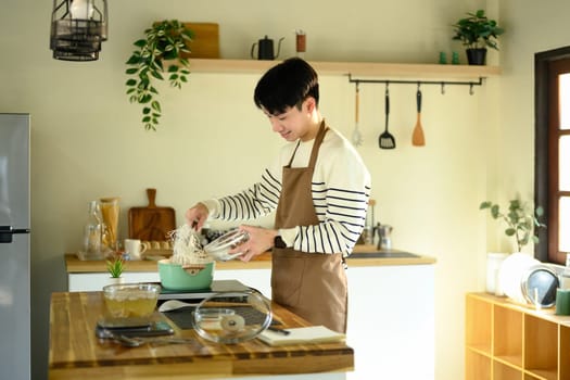 Smiling man in an apron adding noodles to a pot in a sunlit kitchen.