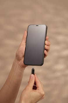 female hand plugging charger cord into smartphone to charge battery on beige background, Wired charging