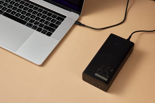 Charging laptop with power bank on beige background, close-up.