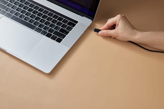 female hand connecting charger cable to laptop at beige table, closeup