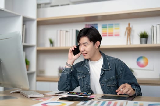 A young designer engaged in a phone call while multitasking at his desk, showcasing effective communication in design.