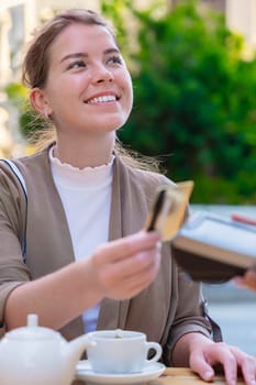 Caucasian woman smiling as she pays with a credit card using contactless technology at an outdoor cafe. A casual urban moment reflecting convenience, lifestyle, and modern financial habits. Vertical