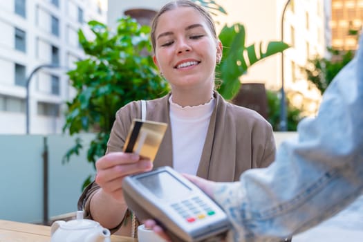 Caucasian woman smiling as she pays with a credit card using contactless technology at an outdoor cafe. A casual urban moment reflecting convenience, lifestyle, and modern financial habits.