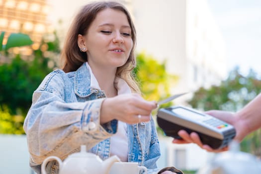 Caucasian woman smiling as she pays with a credit card using contactless technology at an outdoor cafe. A casual urban moment reflecting convenience, lifestyle, and modern financial habits.