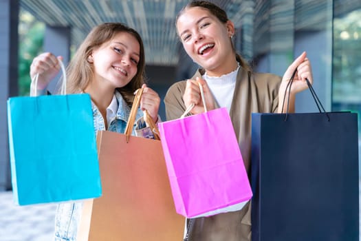 Two cheerful Caucasian women looking at the camera and smiling while holding colorful shopping bags. Capturing joyful and fun shopping moments in an urban outdoor setting.