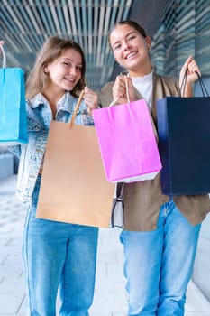 Vertical portrait Two cheerful Caucasian women looking at the camera and smiling while holding colorful shopping bags. Capturing joyful and fun shopping moments in an urban outdoor setting.