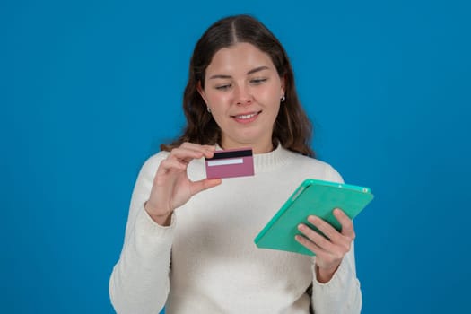 Young woman holding a credit card and a tablet, looking thoughtful.She wears a white sweater and stands against a blue background. Concept of online shopping, payment,or financial decisions.Copy space