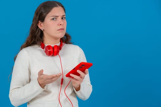 Young woman looking confused and frustrated while holding a red smartphone. Wearing red headphones around her neck, she reacts to a problem or error against a bright blue background. Copy space