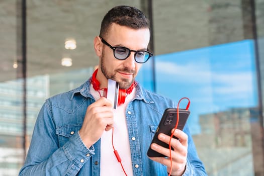 Happy man celebrating a successful online purchase while holding a credit card and smartphone. He smiles with excitement, wearing headphones and sunglasses in a modern urban environment.