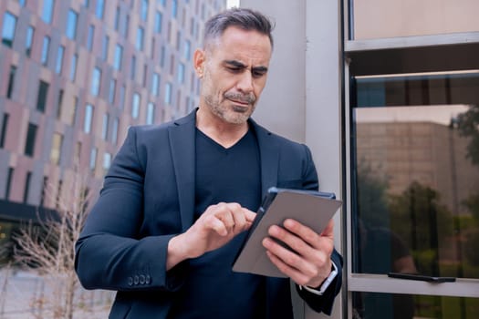 Confident businessman standing outdoors using a tablet in a modern urban setting. He appears focused and professional. Concept of digital work, technology, and contemporary business lifestyle.