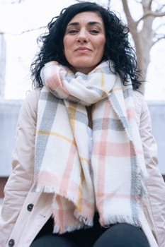 Vertical portrait of a woman sitting on a bench outdoors. She wears a beige coat and scarf, looking calm and confident. Urban background with winter atmosphere and seasonal fashion concept. Vertical