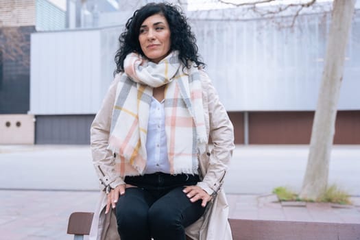 Horizontal portrait of a woman sitting on a bench outdoors. She wears a beige coat and scarf, looking calm and confident. Urban background with winter atmosphere and seasonal fashion concept.
