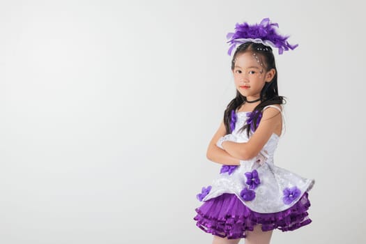 Little girl in a purple feather headpiece, wearing a cheerleader costume with a white and purple layered skirt. She stands confidently with arms crossed. Studio shot on a white background