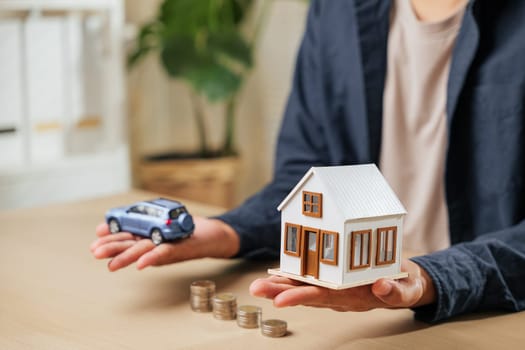 Person displays car and house models above coin stacks, illustrating money-saving strategies for auto and real estate investments. Perfect for business, banking, and personal financial goals.