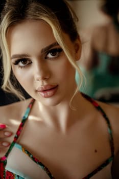 close-up of a stylish blonde in a turquoise dress, in the interior, near the window. the girl has a high hairstyle, bright makeup. on the corset bright red embroidery of flowers. looking at the camera