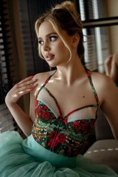 close-up of a stylish blonde in a turquoise dress, in the interior, near the window. the girl has a high hairstyle, bright makeup. on the corset there is bright red embroidery of flowers, dreaming