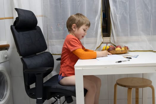 Young boy doing homework, sitting on a big office chair and drawing on a notebook placed on the kitchen table