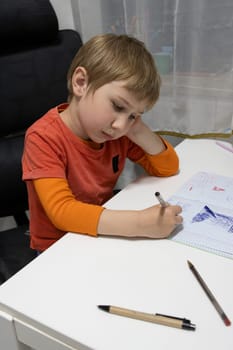 Young boy drawing shapes on a squared notebook, sitting at a white table