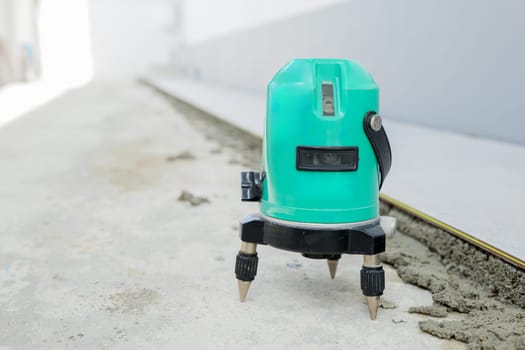 Close-up of a green laser level on a tripod, projecting straight lines to guide the placement of ceramic tiles on a white and gray floor.