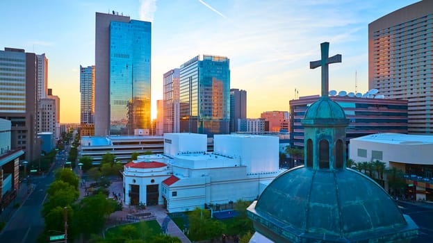 Golden hour aerial view of downtown Phoenix with modern glass skyscrapers glowing in sunset light, a historic church dome with cross in the foreground, and lush city streets below. Urban Southwest vibrancy.