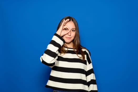Hispanic teenage girl about 15 years old wearing striped sweater over isolated blue background gesturing thumbs up with hand smiling, looking through fingers with happy face