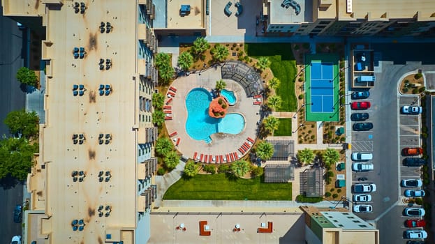 Aerial view of a modern St George Utah apartment complex featuring a resort style swimming pool, lush palm landscaping, and a vibrant blue tennis court under bright midday sun for upscale urban living