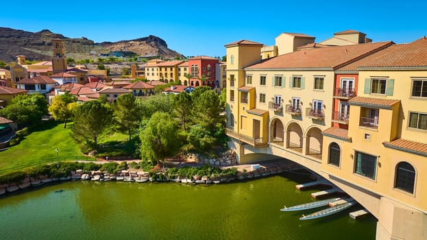 Aerial view of a vibrant Mediterranean-style village with pastel waterfront buildings and rowing boats on Lake Las Vegas, Nevada, set against desert hills and lush green parklands under clear blue skies.