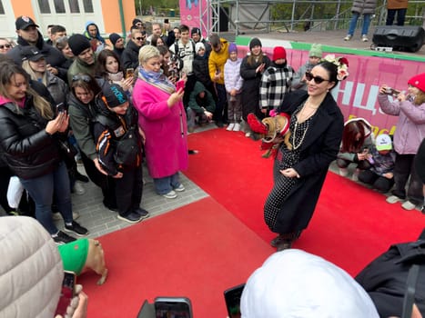 Moscow, Russia, April 27, 2025: Stylish woman walks the red carpet with her dressed up French Bulldog during costume show at the French Bulldog Parade in Vorontsovsky Park, drawing a large crowd