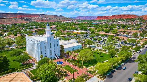 Aerial view of the St George Utah Temple with its striking white architecture surrounded by lush gardens, palm trees, and cityscape set against dramatic red rock cliffs in sunny St George USA