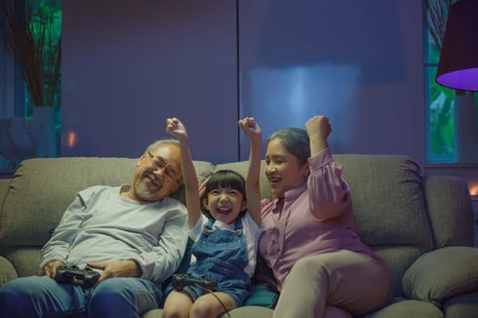 Happy Asian family bonding over a video game on a cozy couch, little girl winning after playing video games console with senior grandparents by controller joysticks while sitting on the sofa