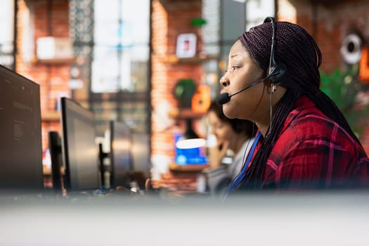 Call center representative in office helping customers by answering questions, close up shot. African american woman in customer support workspace using headset to talk with clients