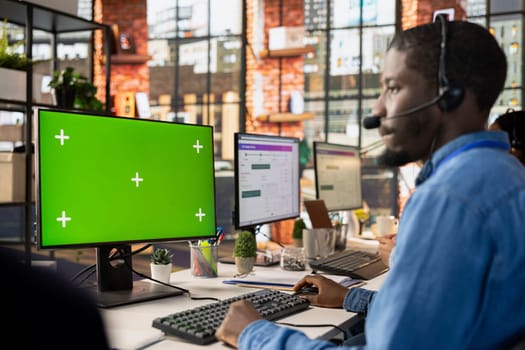 Call center worker in office solving customers issues using green screen PC. African american man in customer support job in front of chroma key computer display using headset to talk with clients