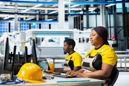 Black woman engineer finetuning energy control systems on computer setup, surrounded by solar power manufacturing. Female expert balances photovoltaics and industrial production.
