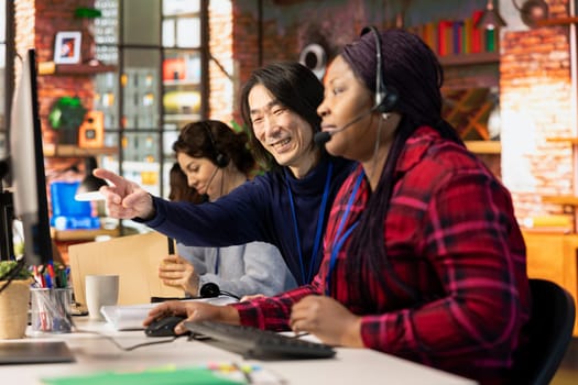Happy customer service senior manager teaching new employee how to track support tickets in company software. Cheerful Asian call center supervisor shows intern how to create and close support tickets