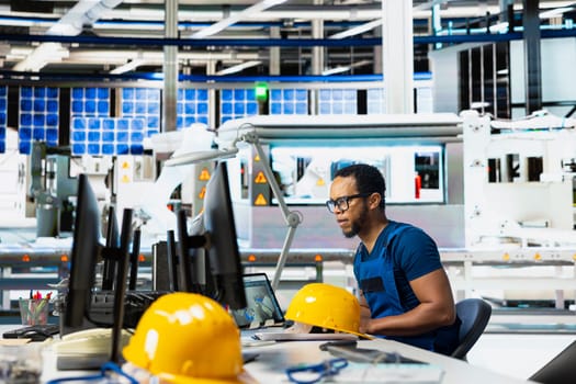 Male engineer using pc setup to oversee solar panel plant assembly lines, troubleshooting photovoltaic system to manage seamless production line with robotic arms. Renewable energy.