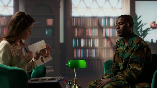 Female therapist presents a Rorschach inkblot test to a black soldier in armed forces uniform during a counseling session. Diverse people at therapy in a warm office with bookshelves. Camera B.