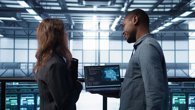 Software developers in data center using laptop, employing AI machine learning models. Workers using notebook to fulfill artificial intelligence infrastructure needs, allocating resources, camera B