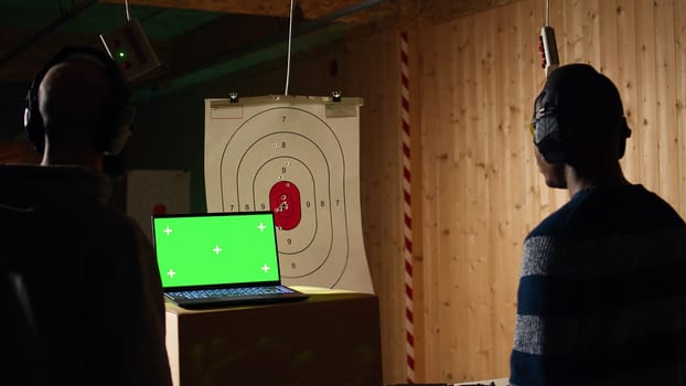 Green screen laptop in shooting range next to man successfully shooting targets. Mockup notebook next to person being congratulated by instructor for accurately hitting bullseye, camera A