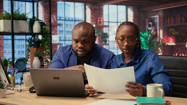 Two remote employees examining calendar appointments on their documents, checking papers and laptop information to solve important tasks on schedule. Couple collaborating at home. Camera A.