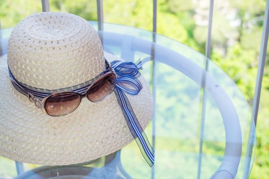 Summer straw hat and sunglasses close-up on a glass table on the balcony.